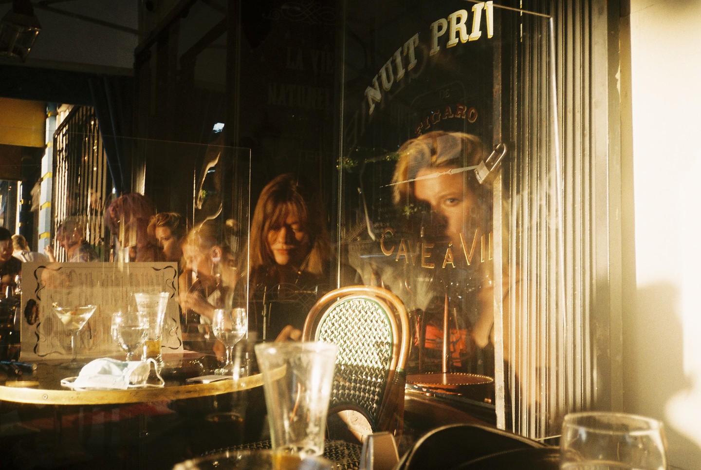 A woman looks out from a cafe window with reflections of other patrons and the street.