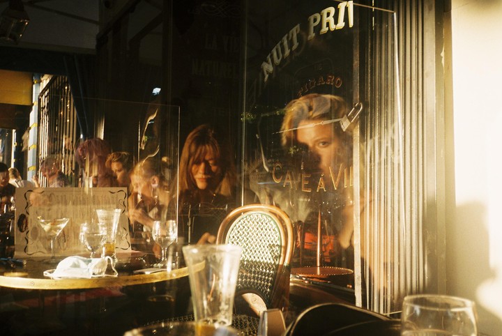 A woman looks out from a cafe window with reflections of other patrons and the street.