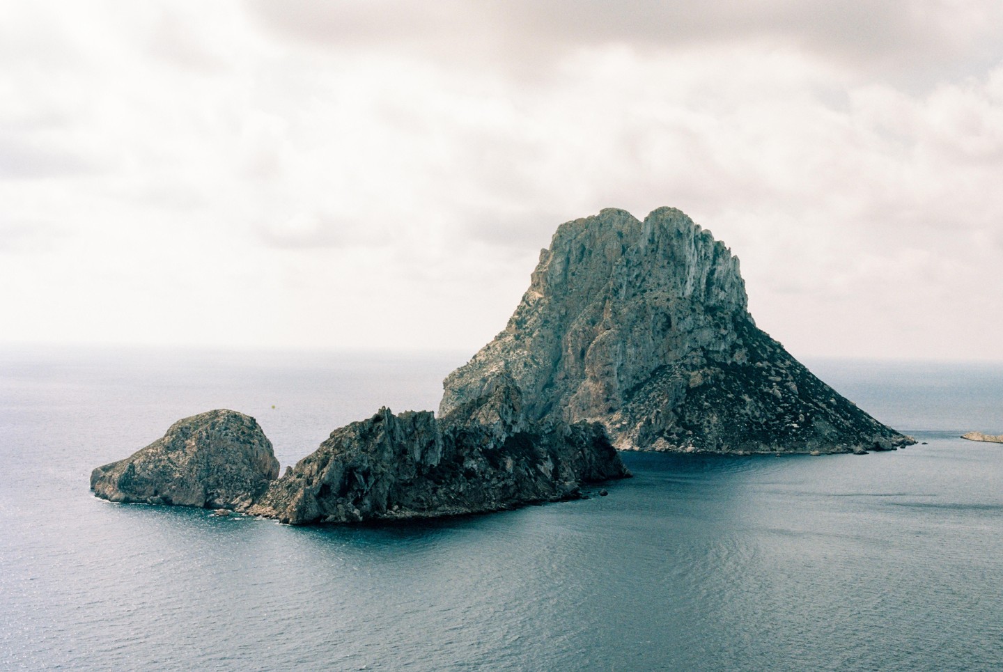 A large, rocky island rises from the sea under a cloudy sky, with smaller rock formations in the foreground.