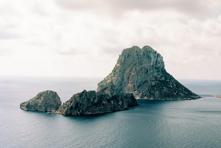 A large, rocky island rises from the sea under a cloudy sky, with smaller rock formations in the foreground.