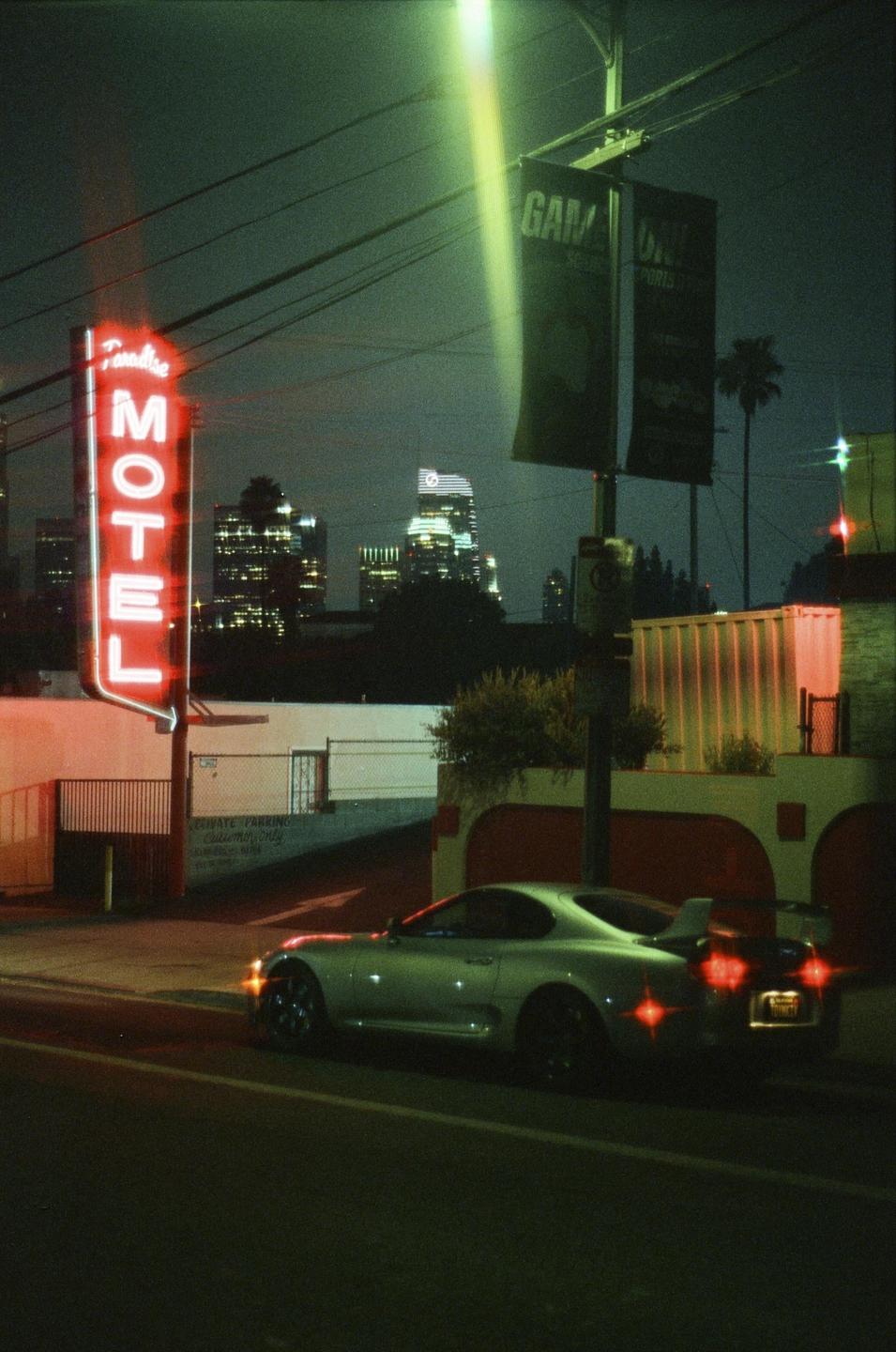 A silver sports car is parked on a street at night, with a neon motel sign and city lights in the background.