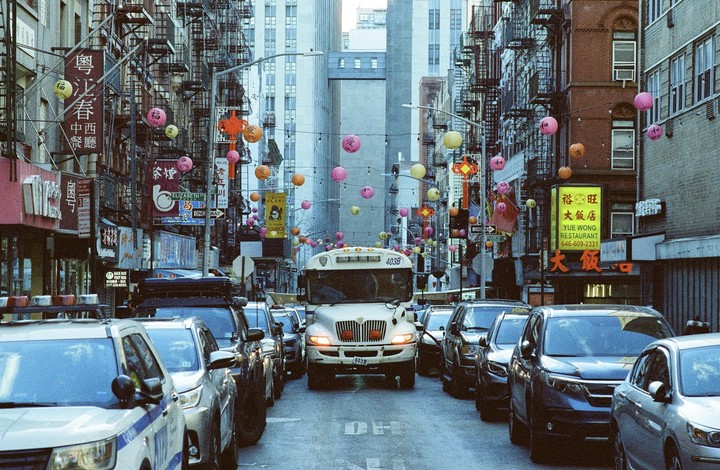 A white bus is stuck in traffic on a narrow street decorated with colorful lanterns in Chinatown, New York.