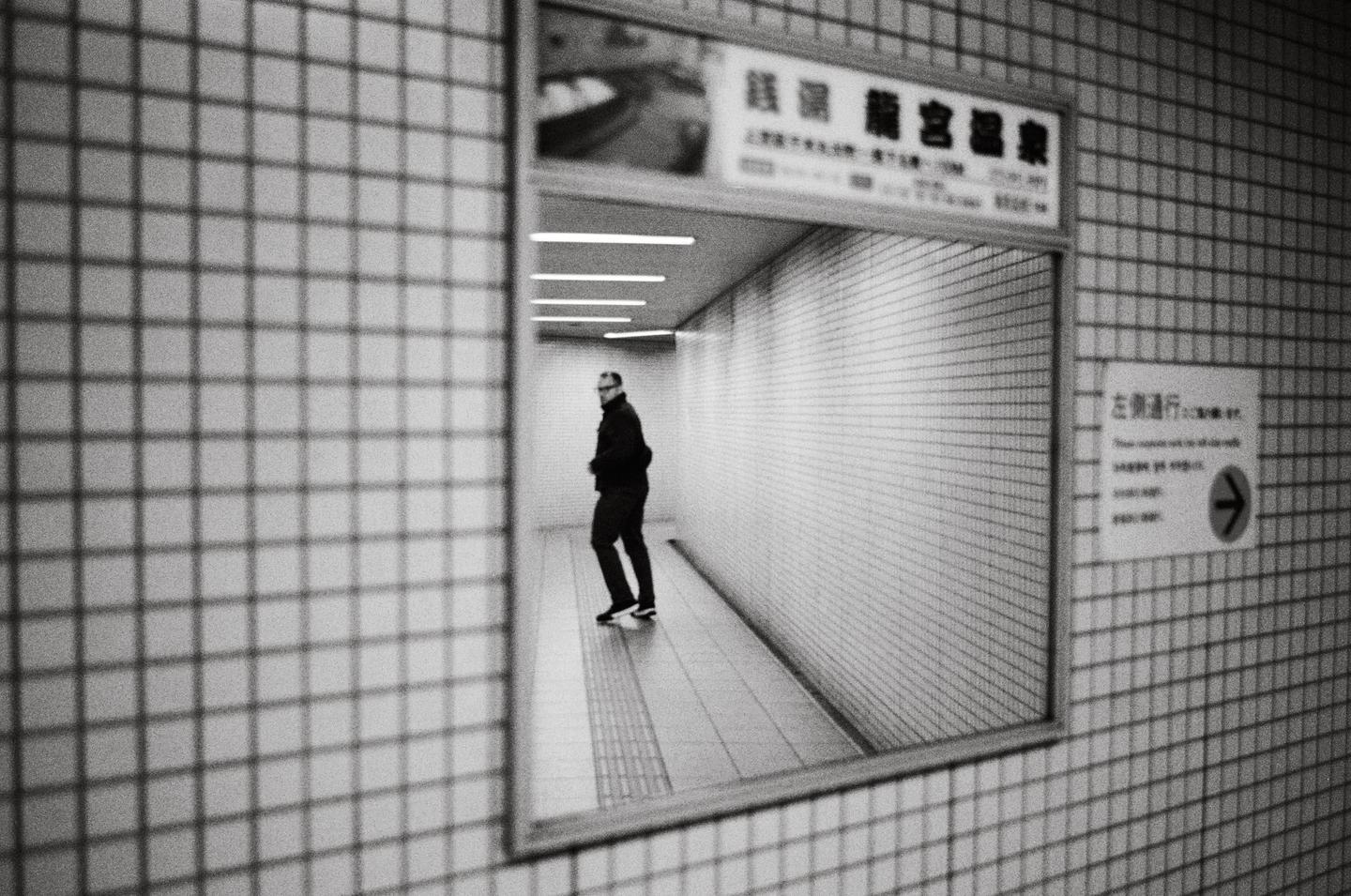 A man walks down a tiled hallway, reflected in a mirror on a tiled wall.