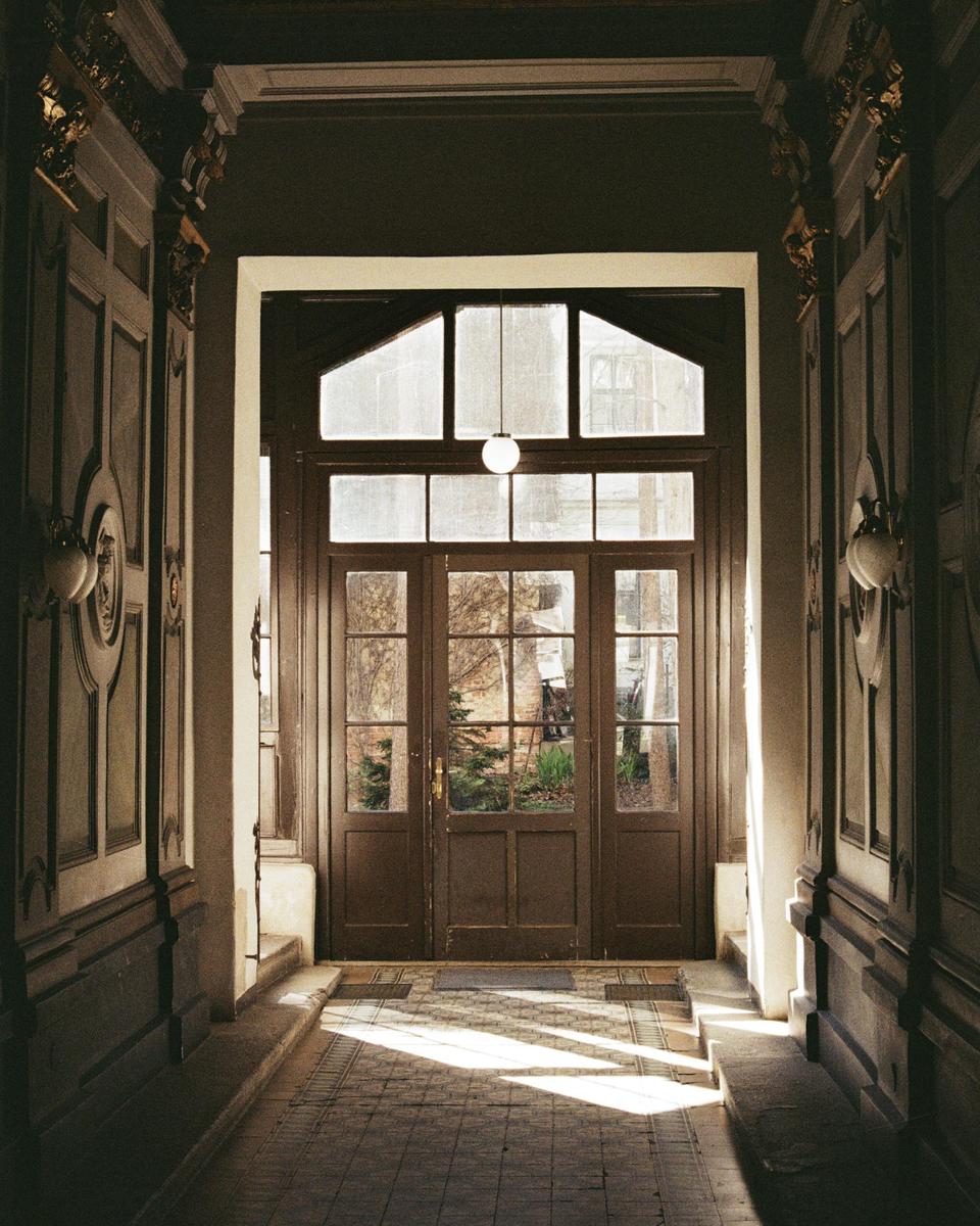 Sunlight streams through a glass door at the end of a long, ornate hallway.