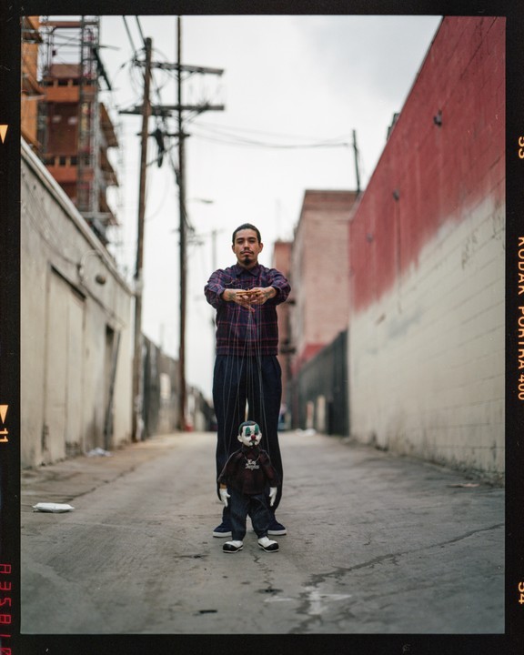 A man stands in an alley holding the strings of a marionette dressed as a clown.