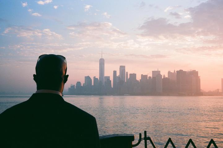 A man in a suit looks out over the Hudson River at the New York City skyline during sunrise.