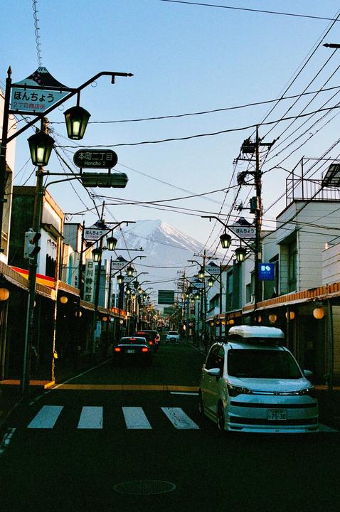A street in Japan is lined with buildings and streetlights, with a snow-capped mountain visible in the distance.