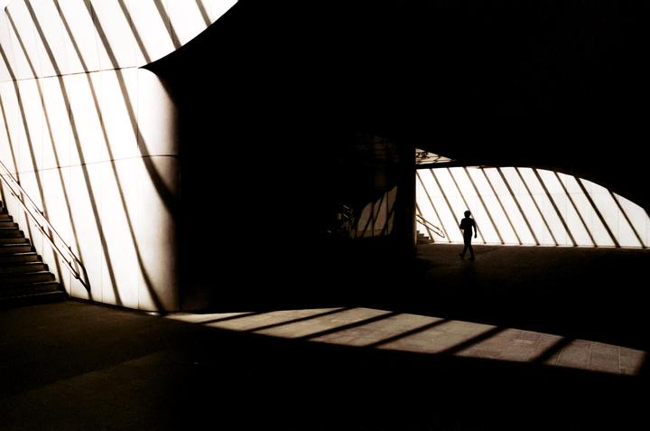 A lone figure walks through a dark, angular space illuminated by strong diagonal shadows.