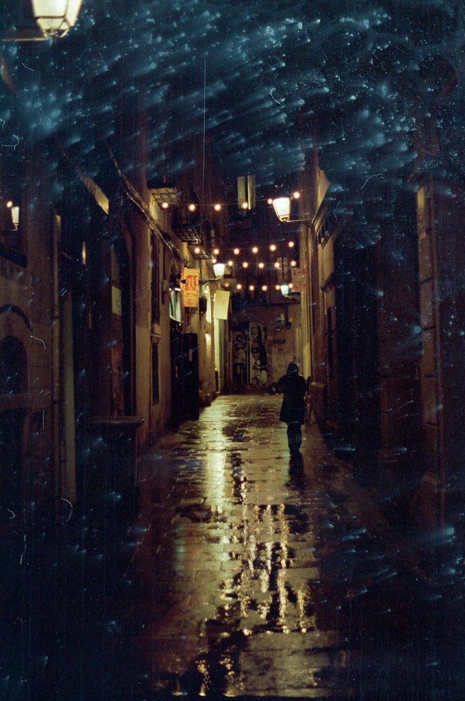 A person walks down a narrow, wet street at night, illuminated by string lights and street lamps.