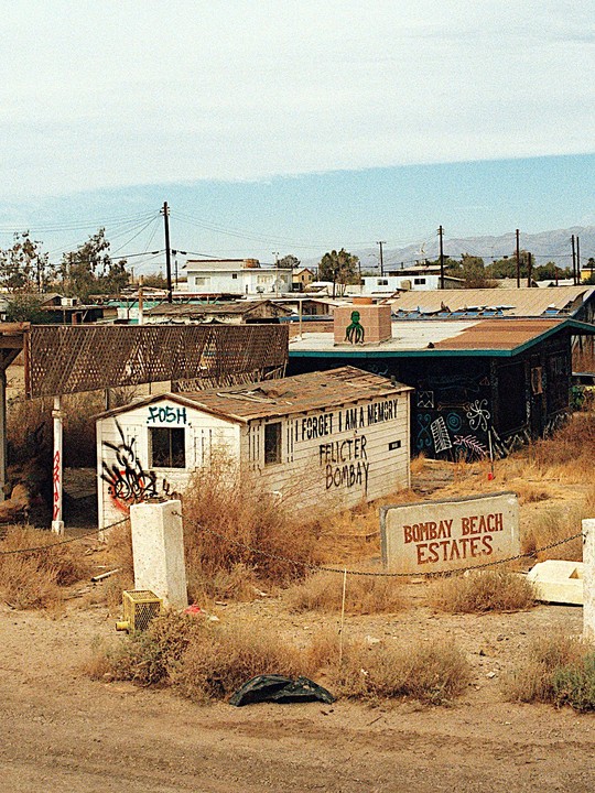 A collection of dilapidated buildings with graffiti and a sign for Bombay Beach Estates are visible under a hazy sky.