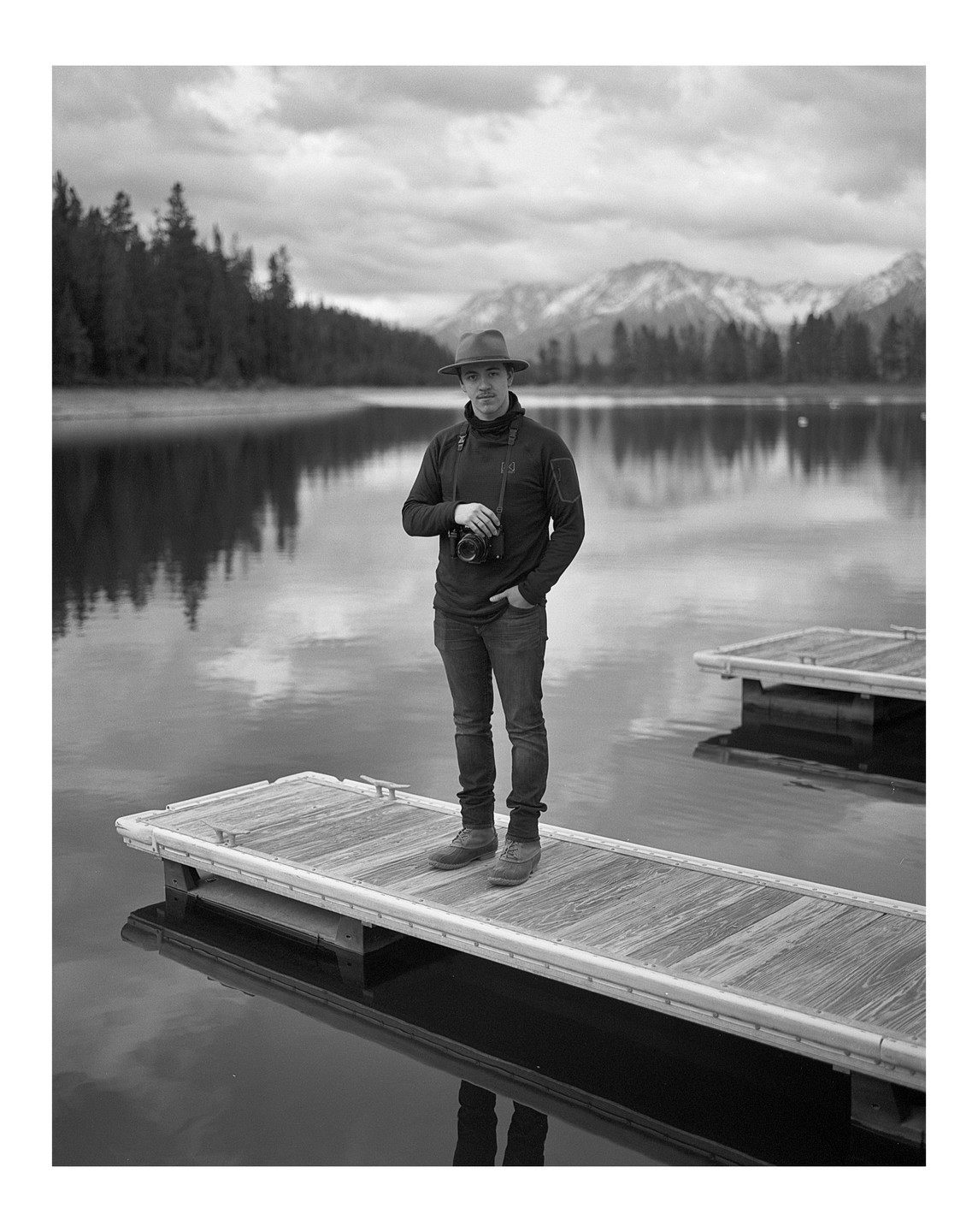 A man wearing a hat and holding a camera stands on a dock in front of a lake and mountains.