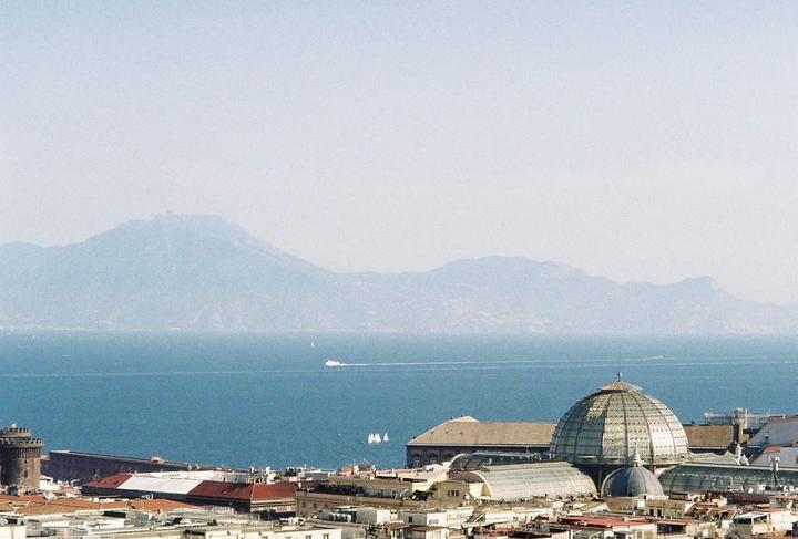 A city with a large domed building in the foreground overlooks a body of water with mountains in the distance.