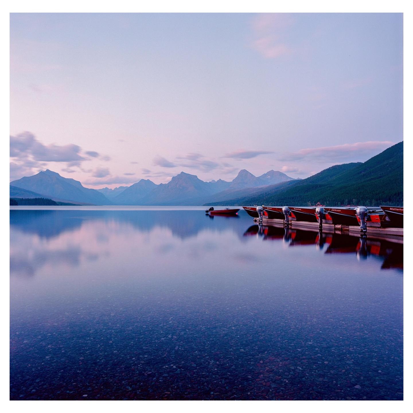 Several red boats are docked on a calm lake with mountains in the background under a soft, colorful sky.