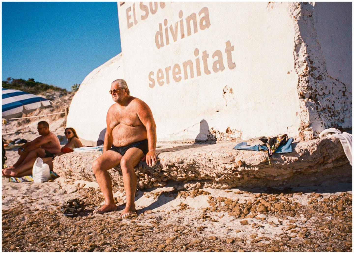 A man in swim trunks sits on a rock wall next to a building with text on it, with other people and a beach umbrella in the background.