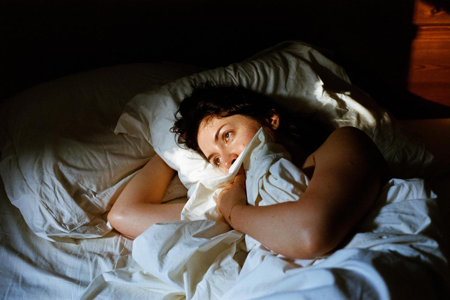 A woman lies in bed, partially covered by a white sheet, with light illuminating her face.