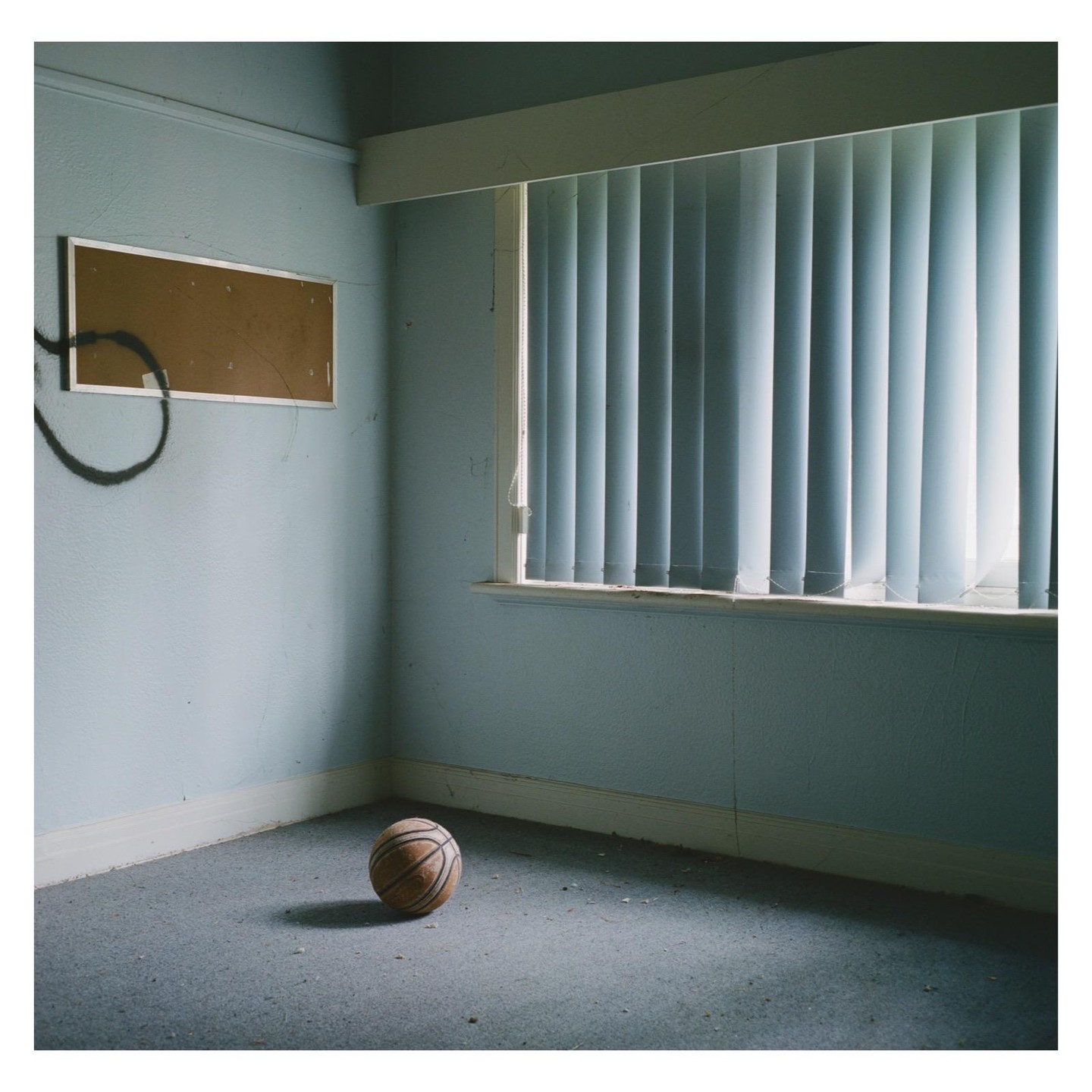 A basketball sits on a dusty carpeted floor in a room with light blue walls and a window with vertical blinds.