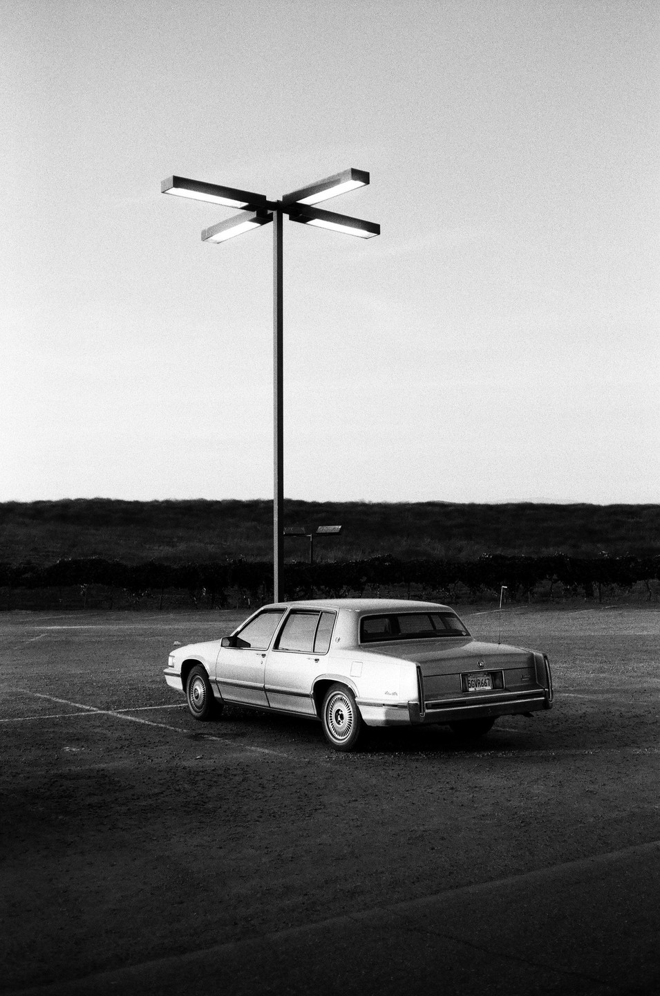 A car is parked in a dirt lot next to a tall light pole with a dark field in the background.