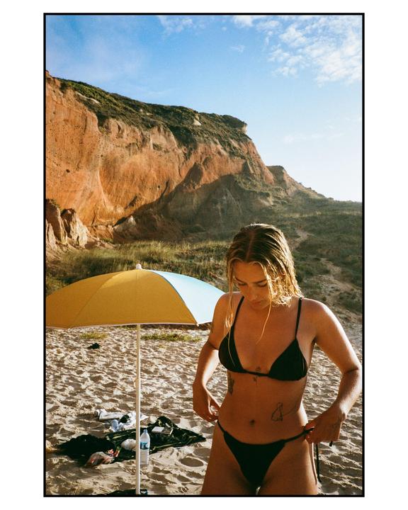 A woman in a black bikini adjusts her swimsuit on a sandy beach next to a yellow and blue umbrella, with a large cliff in the background.
