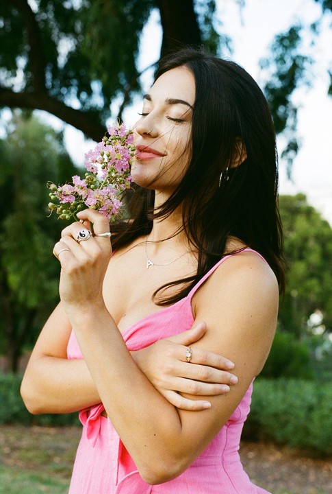 A woman in a pink dress holds a cluster of pink flowers to her nose, with her eyes closed and a slight smile.