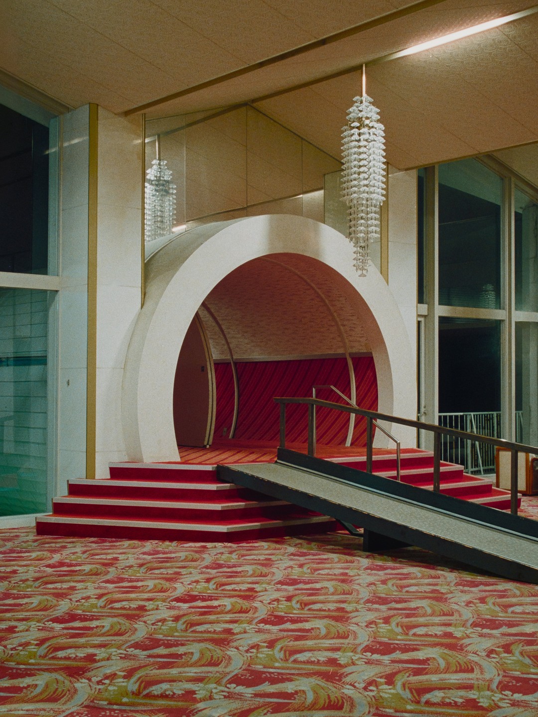 A red carpeted staircase leads to an arched entryway with a ramp beside it, all within a hotel lobby.