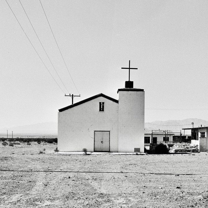 A white church with a cross on its steeple stands in a barren, flat landscape under a bright sky.