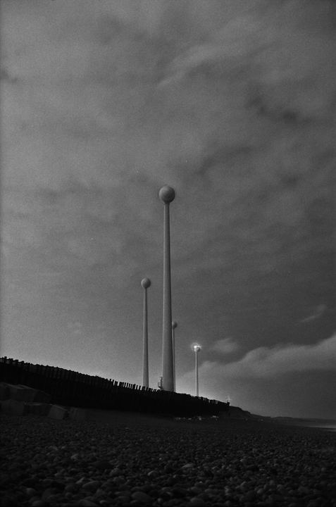 A black and white image shows tall, spherical streetlights on a pebble beach under a cloudy sky.