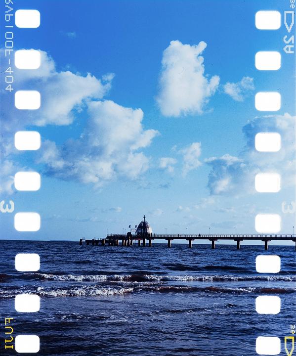 A long pier with a domed building at its end extends into the ocean under a blue sky with white clouds.