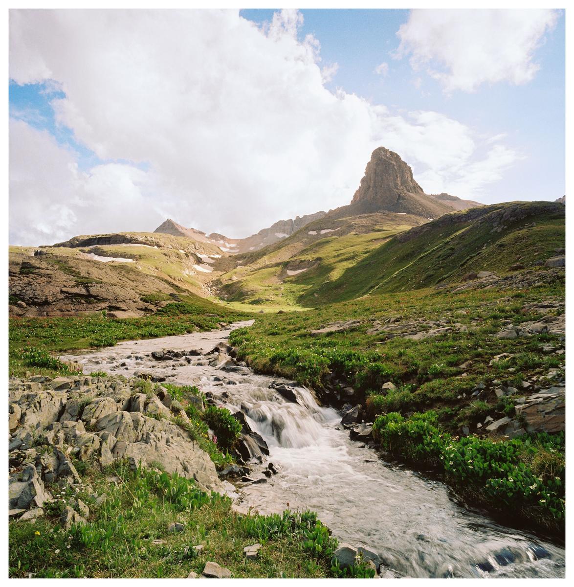 A stream flows through a green valley with mountains under a cloudy sky.