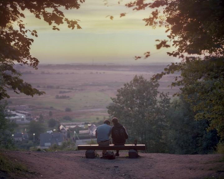 Two men sit on a bench overlooking a valley with a town and fields at dusk.