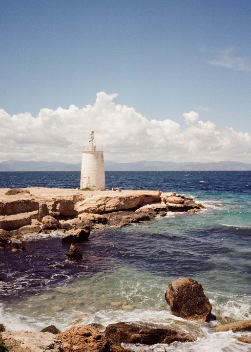 A white lighthouse stands on a rocky shore next to the sea under a blue sky with clouds.