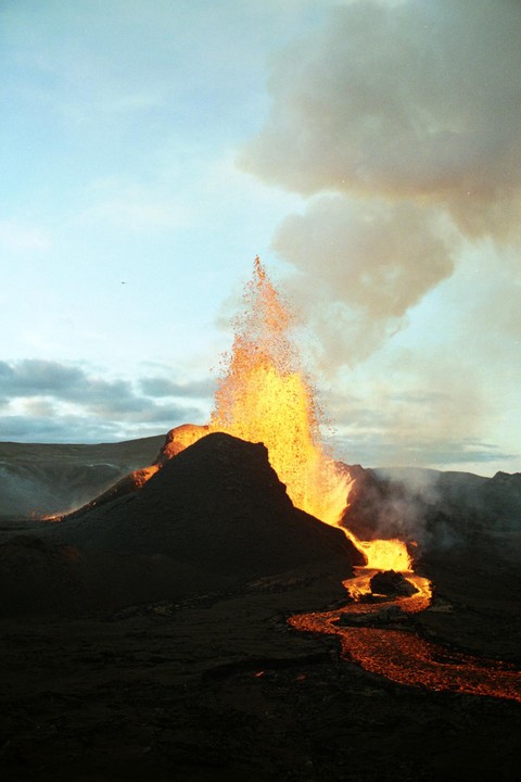 A volcano erupts, spewing lava into the air and down a winding path across the landscape under a cloudy sky.