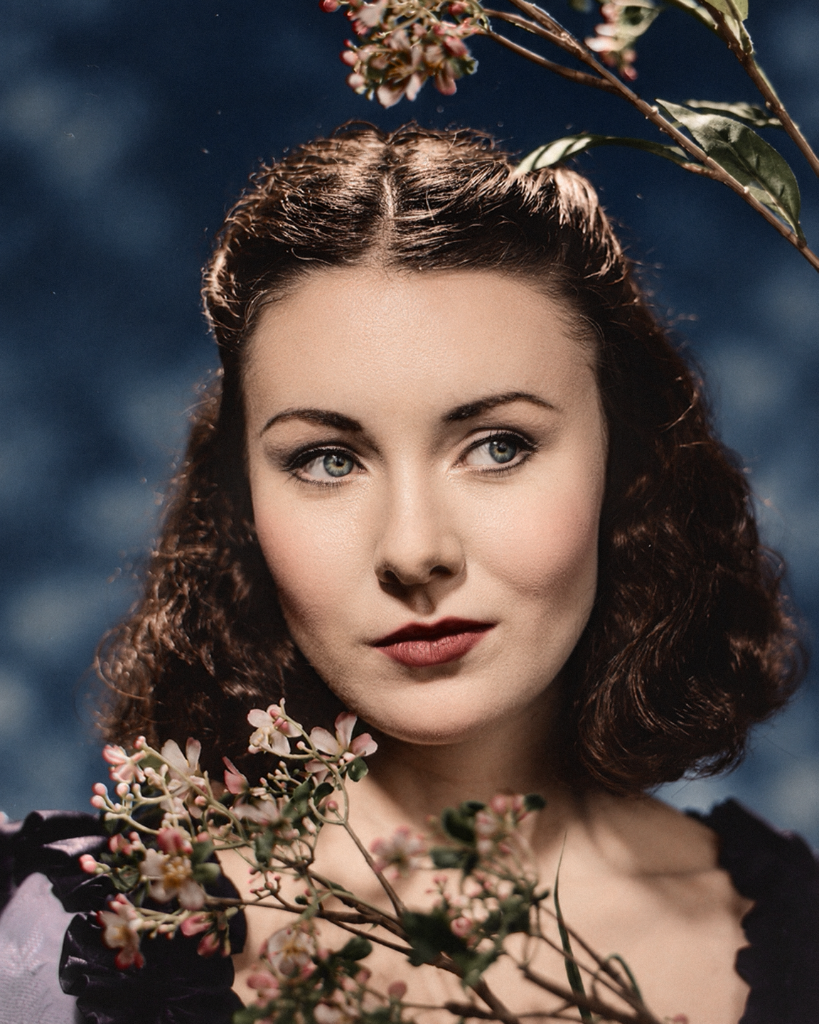 A woman with dark curly hair and red lipstick looks to the side, framed by flowering branches.