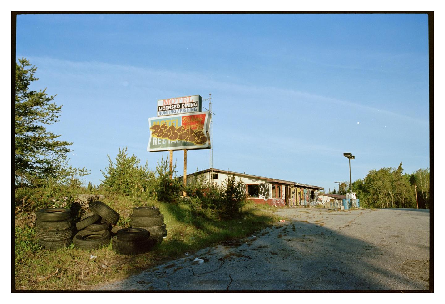 An abandoned motel with a large sign and piles of tires sits beside a cracked asphalt road.