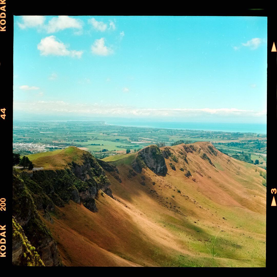 A vast landscape shows rolling hills and a distant coastline under a bright blue sky.