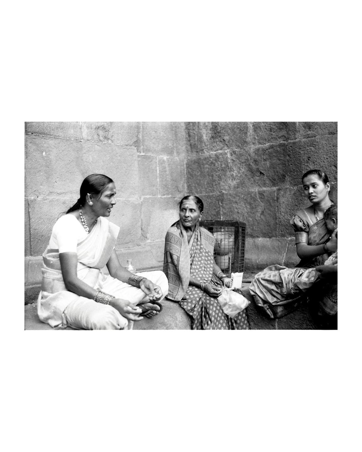 Three women sit on a stone ledge in front of a stone wall, with one woman holding a child.