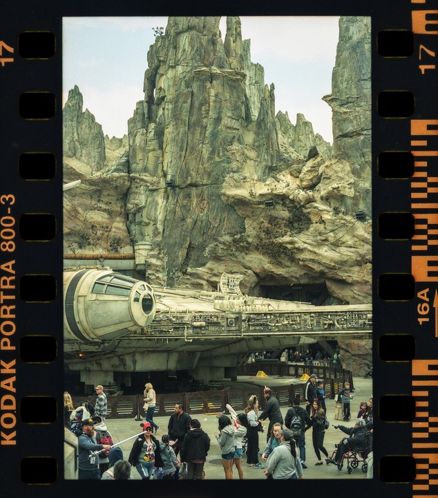 A large spaceship is docked in front of a rocky mountain with many people walking around it.