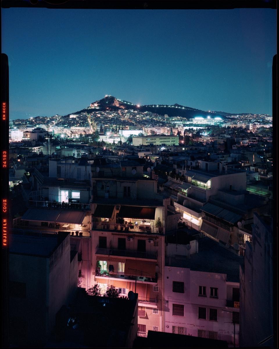 A city at night is illuminated by numerous lights, with a prominent hill in the background also dotted with lights.