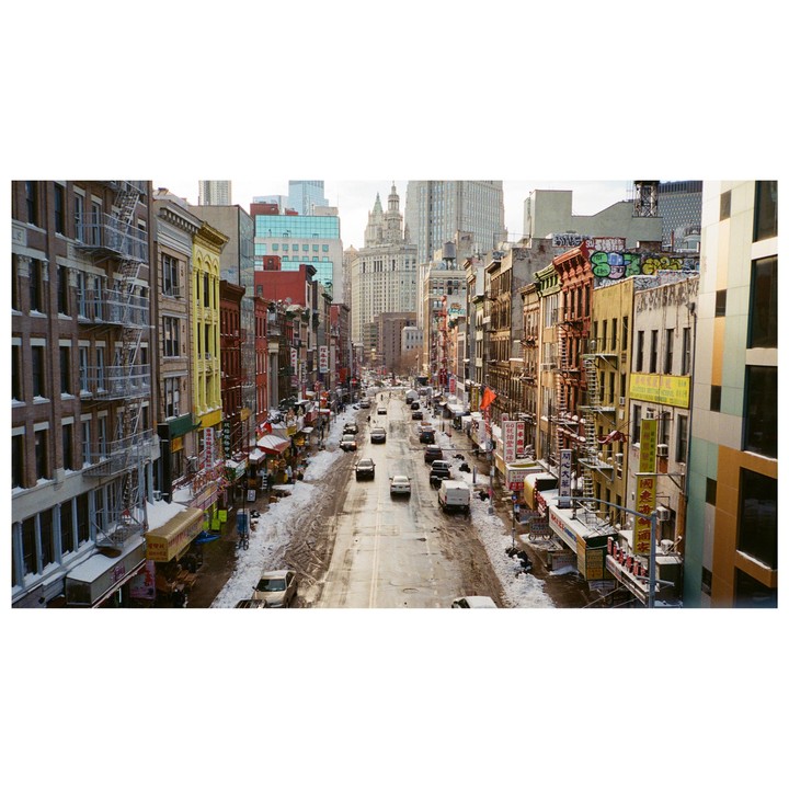 A wide street in a city is lined with colorful buildings and cars, with snow on the sidewalks and a large building in the distance.