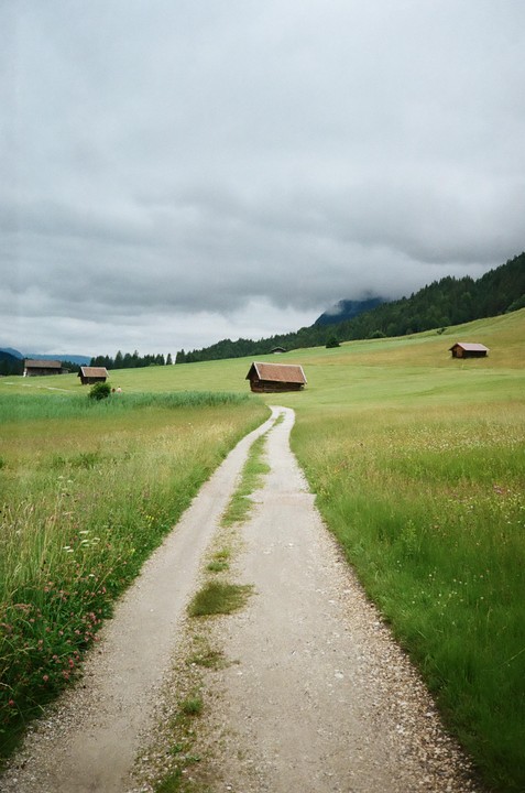 A dirt path winds through a green meadow with several small wooden huts under a cloudy sky.
