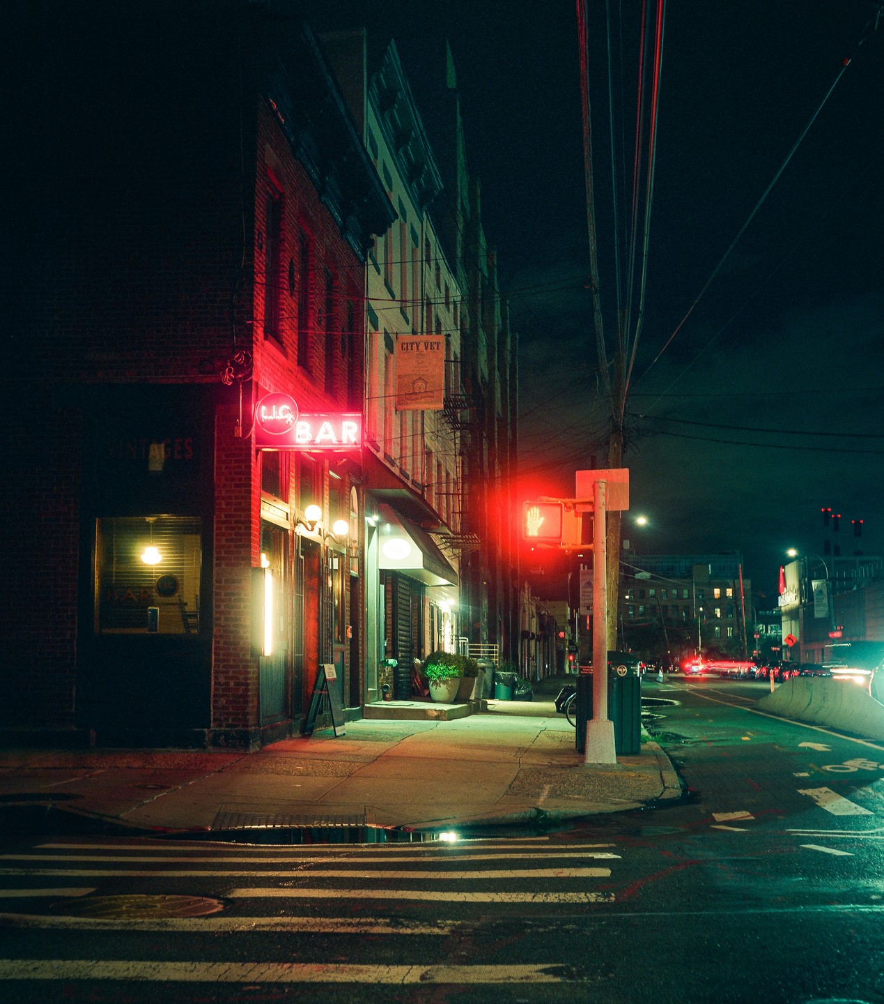 A neon "BAR" sign glows red on a brick building next to a street with a red traffic light and a puddle reflecting the light.