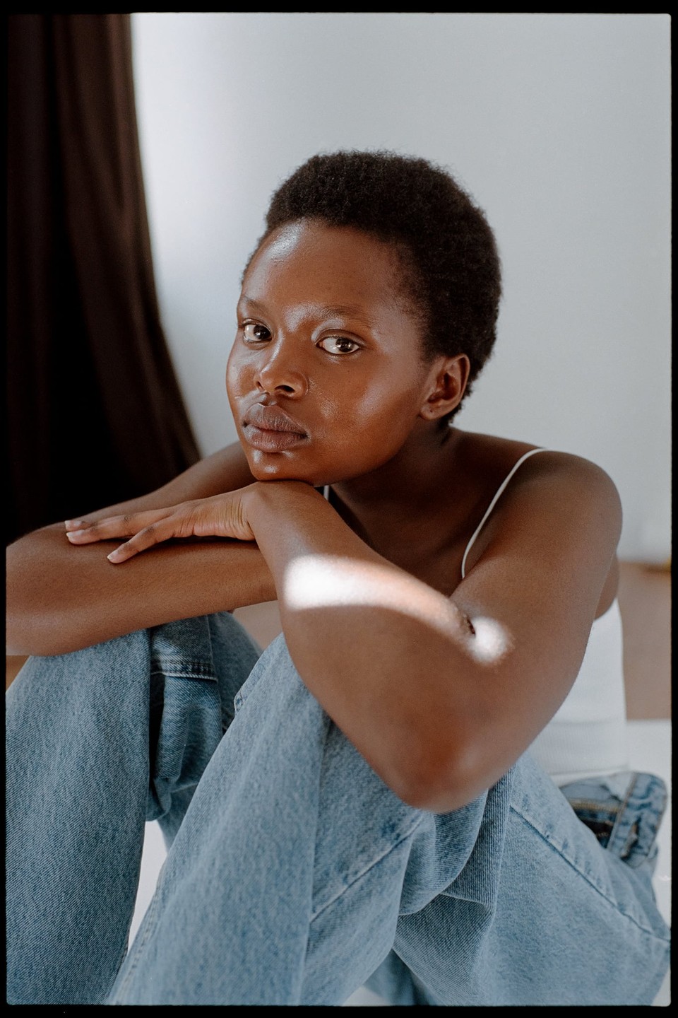 A woman with short hair and light blue jeans sits on the floor with her arms crossed, looking directly at the viewer.