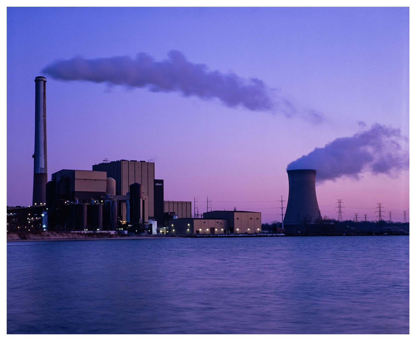 A power plant with two smokestacks emits smoke into the purple dusk sky over Lake Michigan.