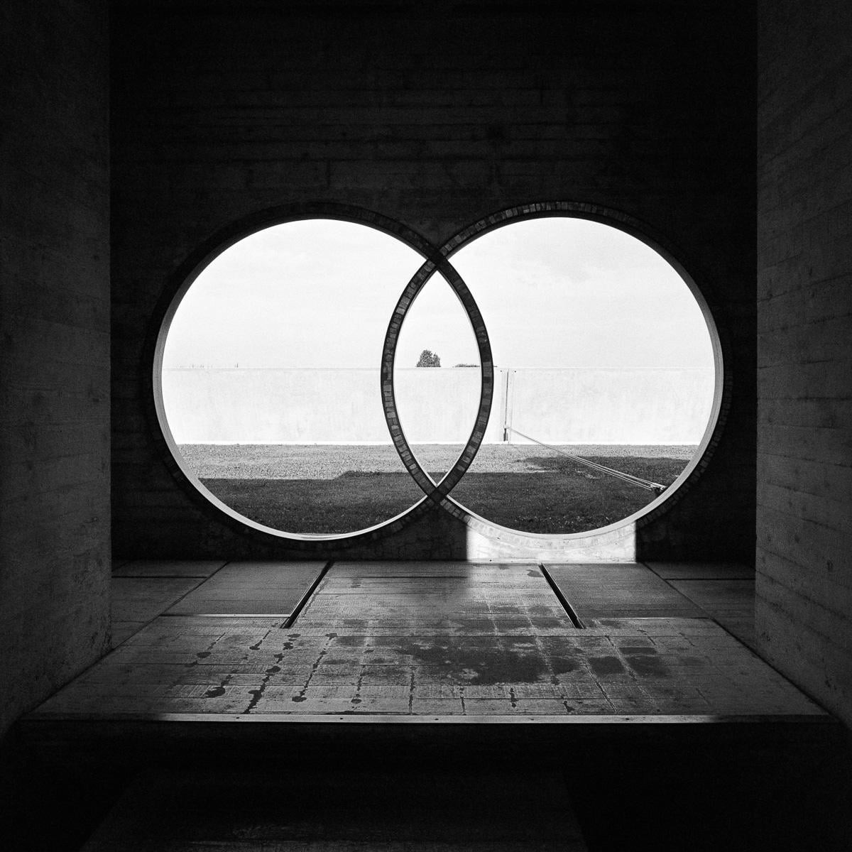 Two large interlocking circular windows frame a distant island in a body of water, seen from inside a concrete structure.