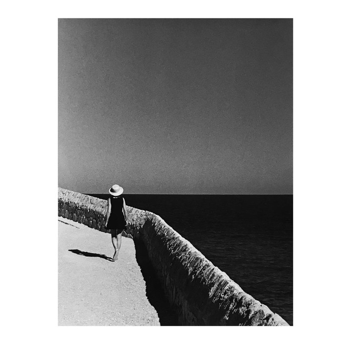 A woman in a hat walks along a stone wall next to the ocean.