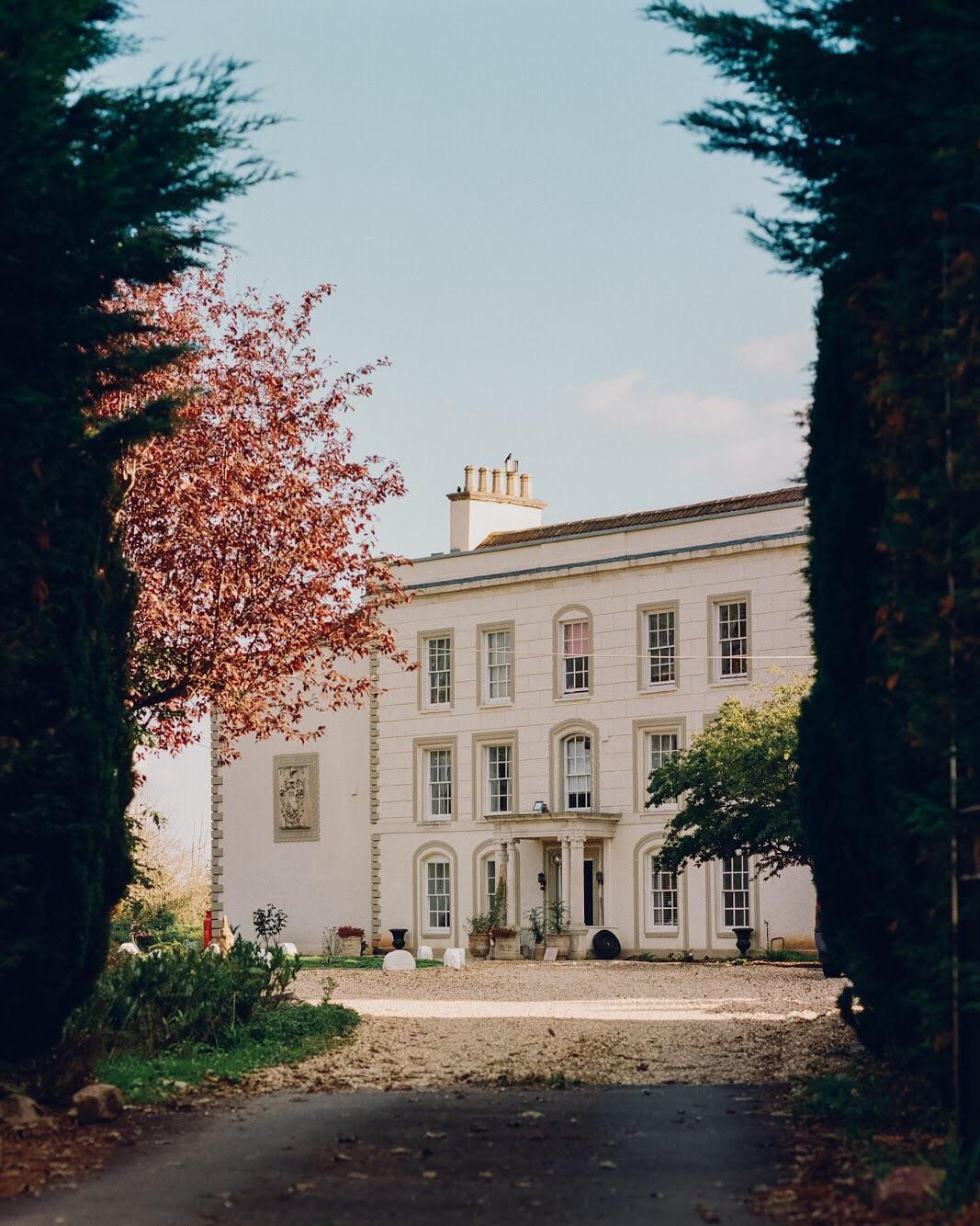 A large white manor house with many windows is seen through an opening in tall trees.
