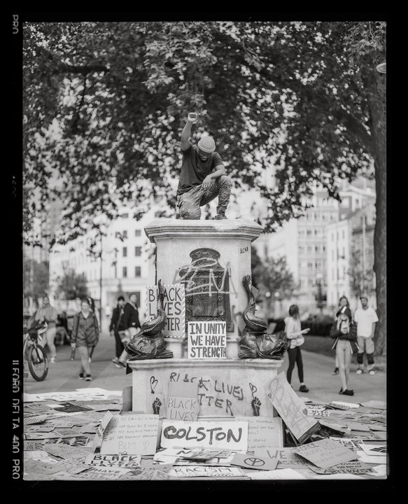 A man kneels on a statue base with his fist raised, surrounded by protest signs.