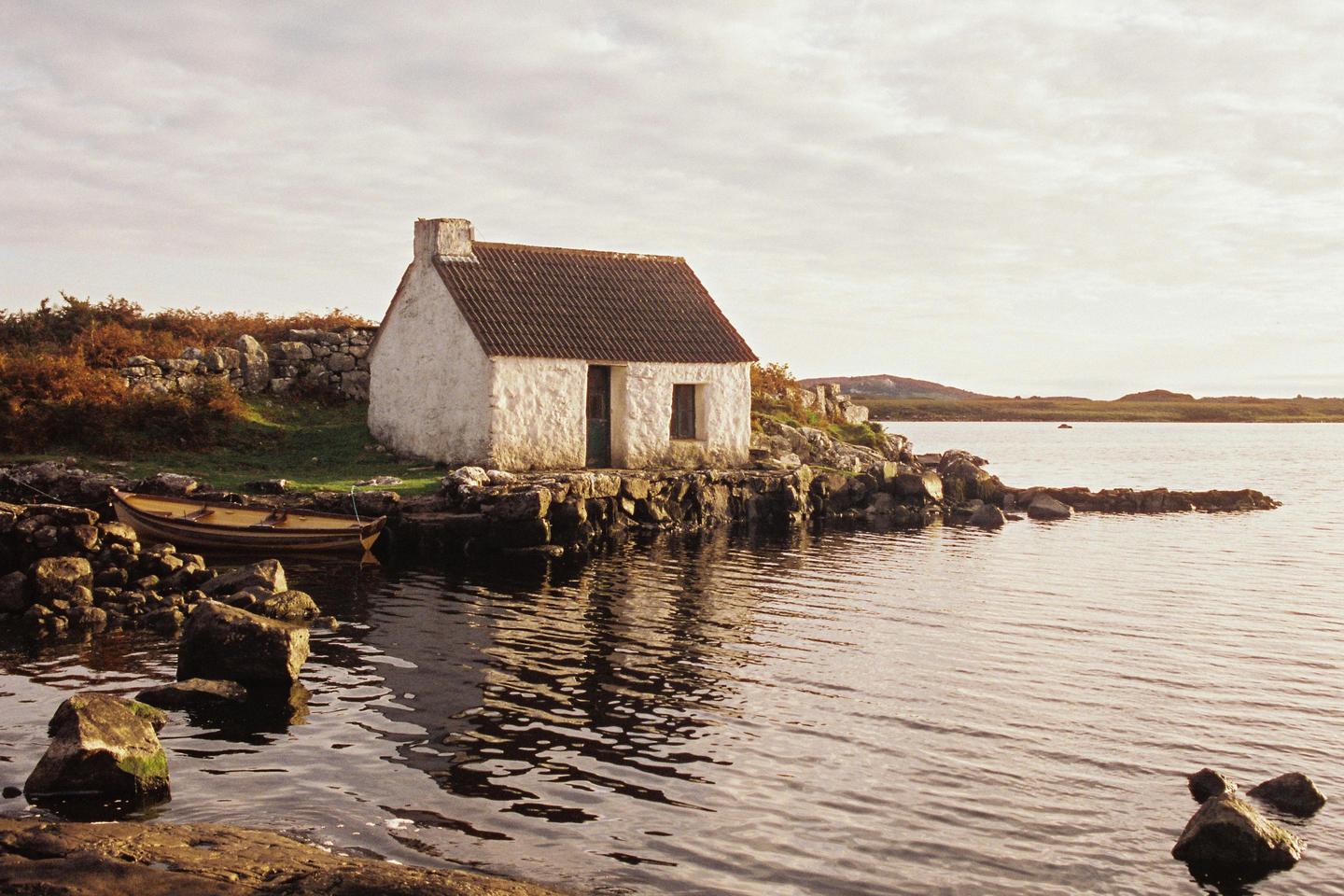 A small white cottage with a brown roof sits on a rocky shore next to a body of water, with a wooden boat moored nearby.