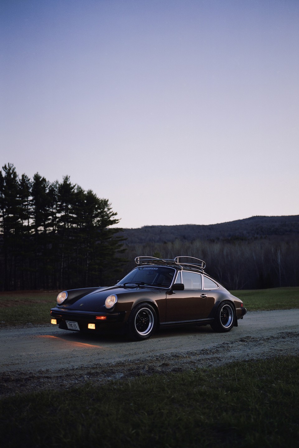 A brown car with a roof rack is parked on a dirt road with mountains and trees in the background at dusk.