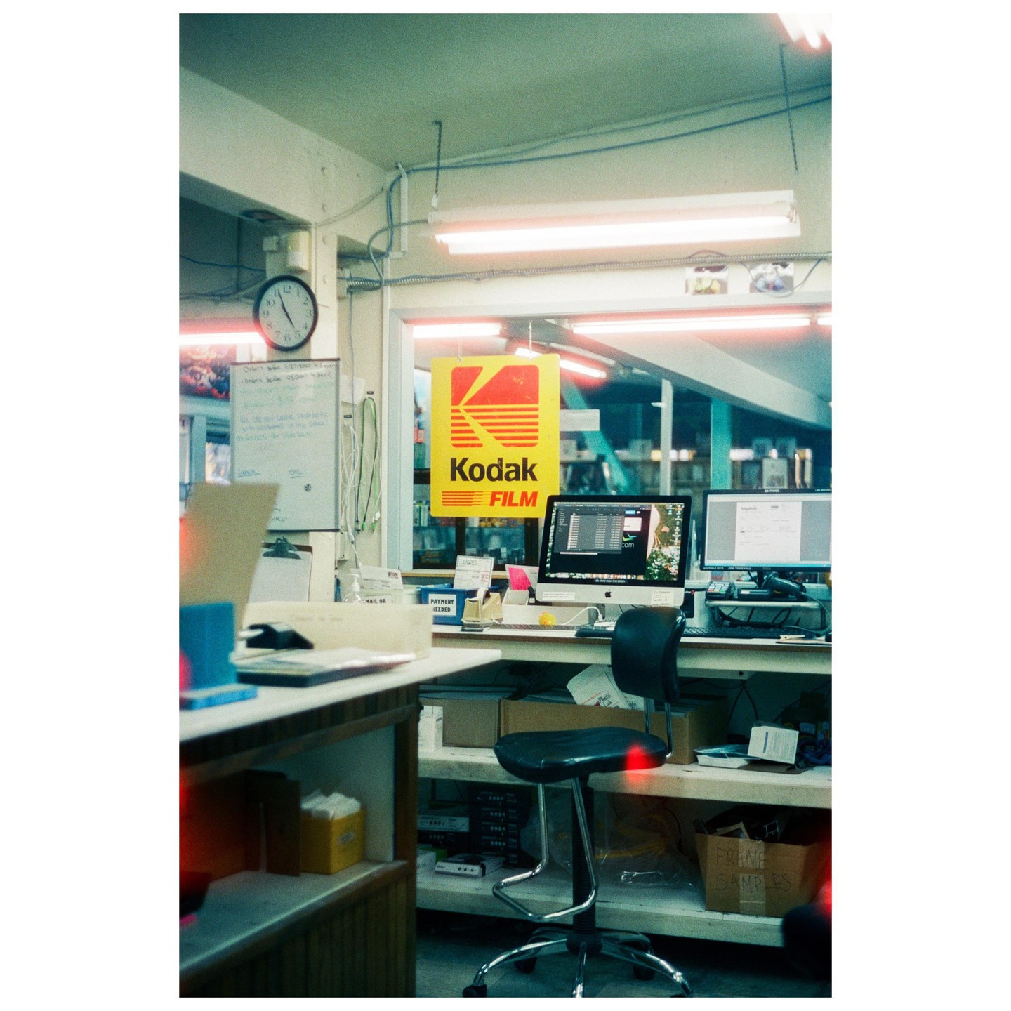A Kodak Film sign hangs above a desk with two computer monitors and a stool in a room with fluorescent lighting.