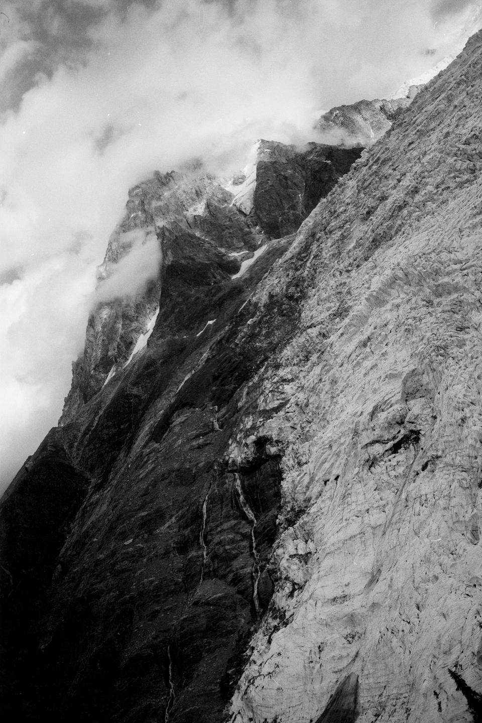 Clouds partially obscure a rugged mountain peak and a glacier in the French Alps.